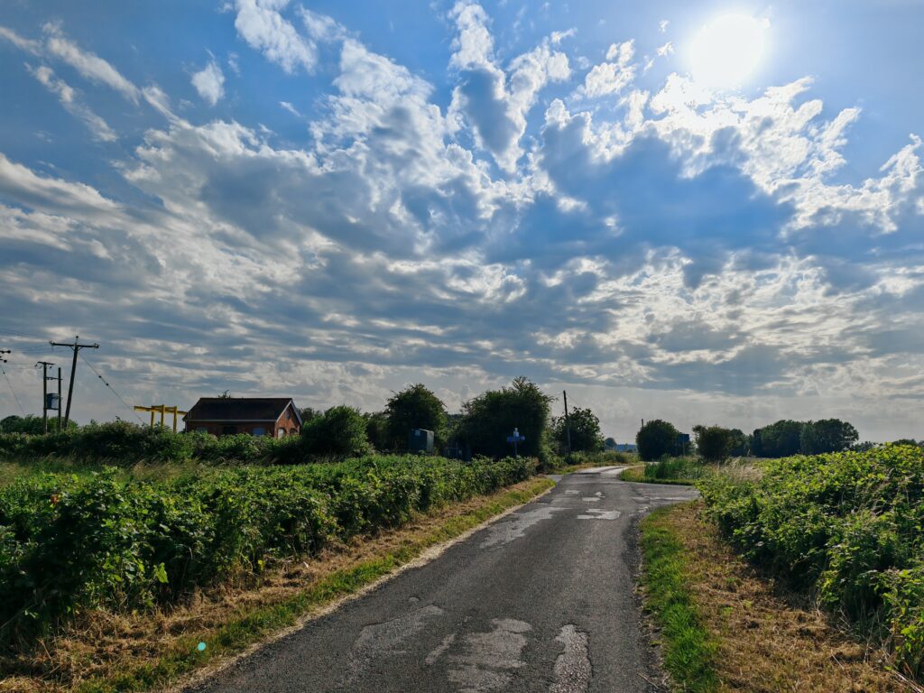 Rural road under a bright, cloudy sky with a red-brick house and lush greenery on either side, bathed in sunlight.