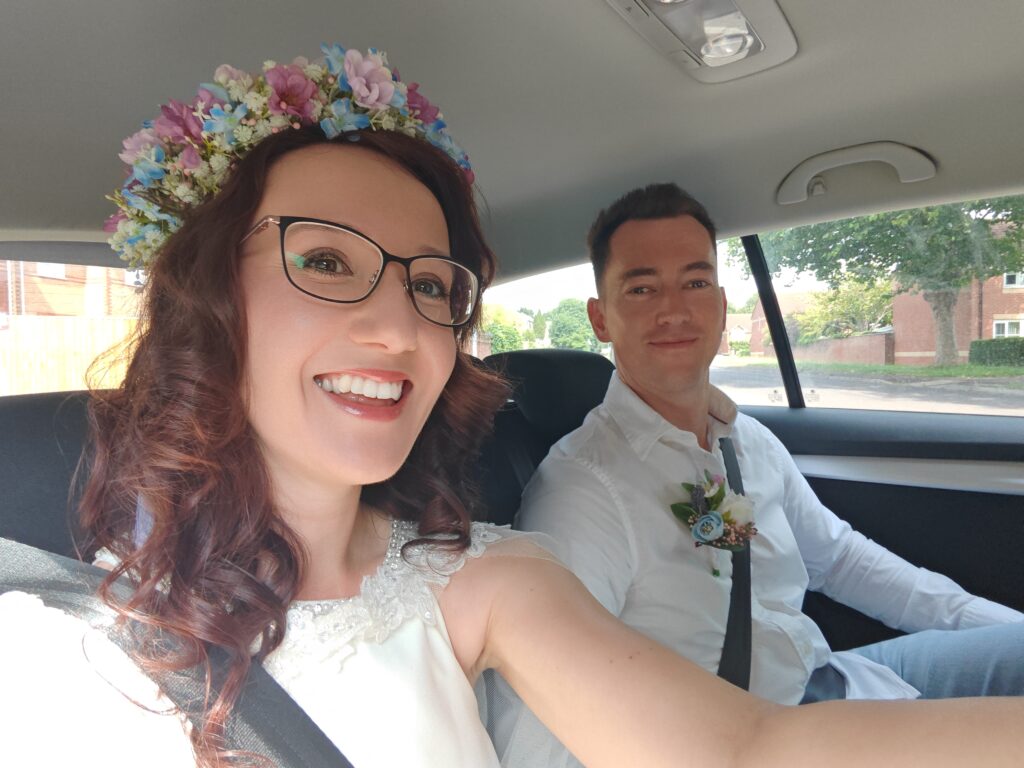 Bride and groom smiling in a car, with the bride wearing a floral crown and the groom in a white shirt.
