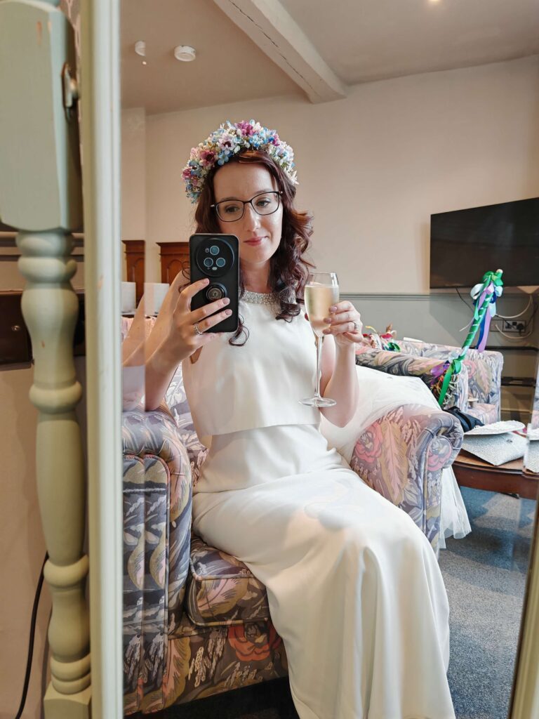 Bride in a white gown and flower crown takes a mirror selfie, holding a glass of champagne, seated on a floral couch.