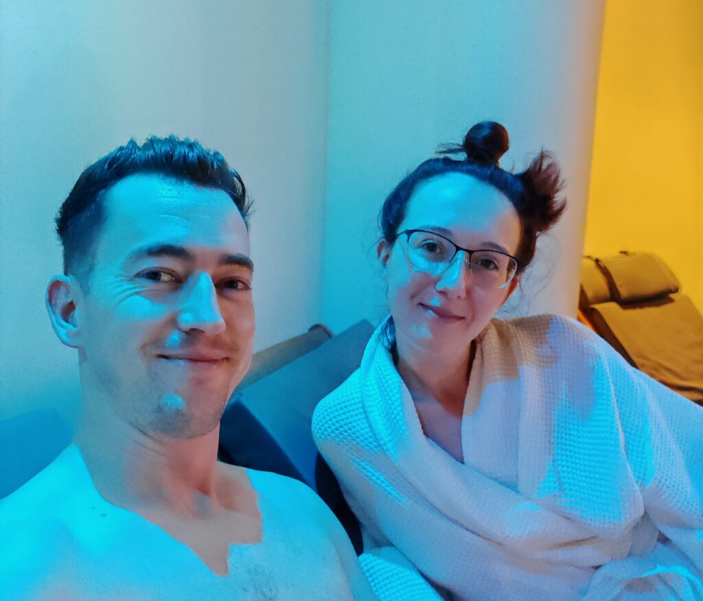 Couple relaxing in a cozy setting with colorful lighting, wearing comfortable robes, smiling at the camera.