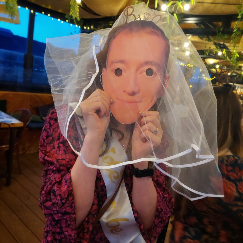 Woman in party attire wearing a humorous face mask and veil at a celebration, holding up mask playfully.
