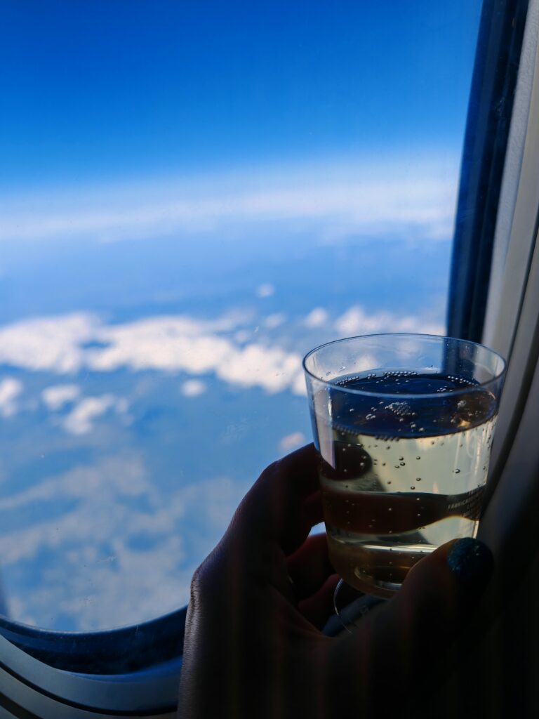 Hand holding a glass of champagne by an aeroplane window, with a clear blue sky and clouds visible outside.