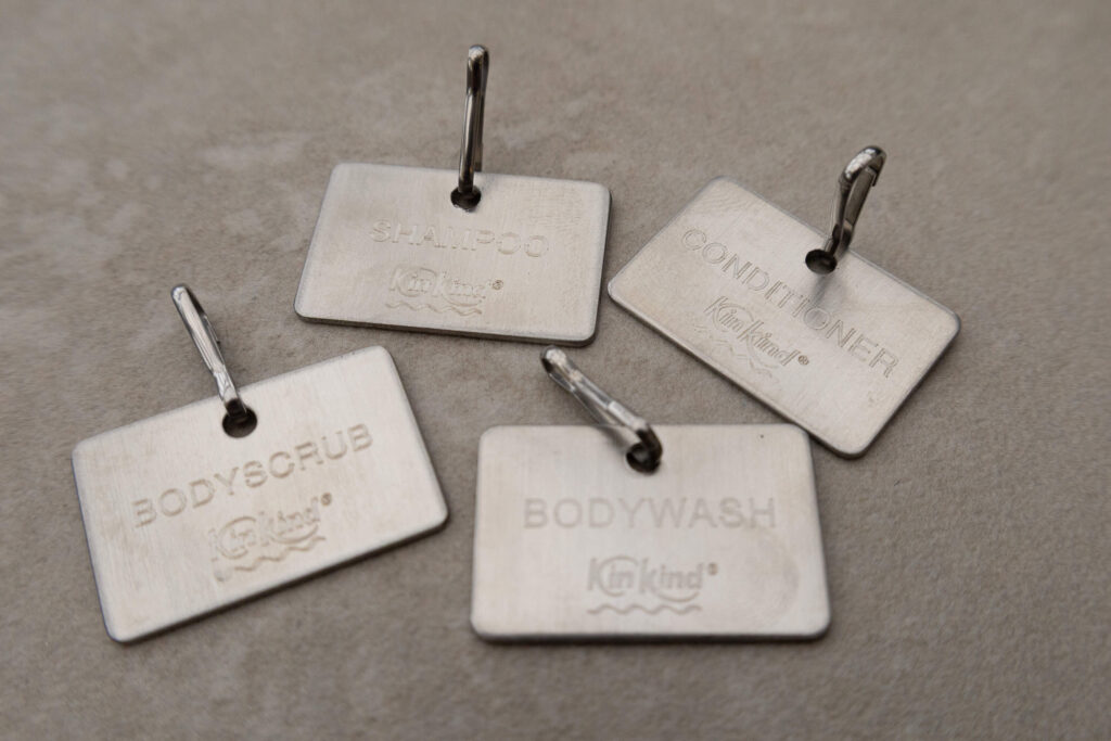 Metal shower caddies with engraved labels for shampoo, conditioner, bodyscrub, and bodywash on a grey background.