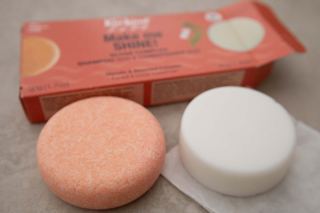 Two solid shampoo bars, one orange and one white, with packaging in the background on a beige surface.