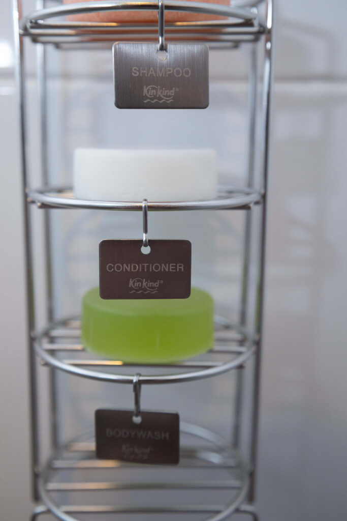 Shampoo, conditioner, and body wash bars on a metal rack with label tags in a bathroom setting.