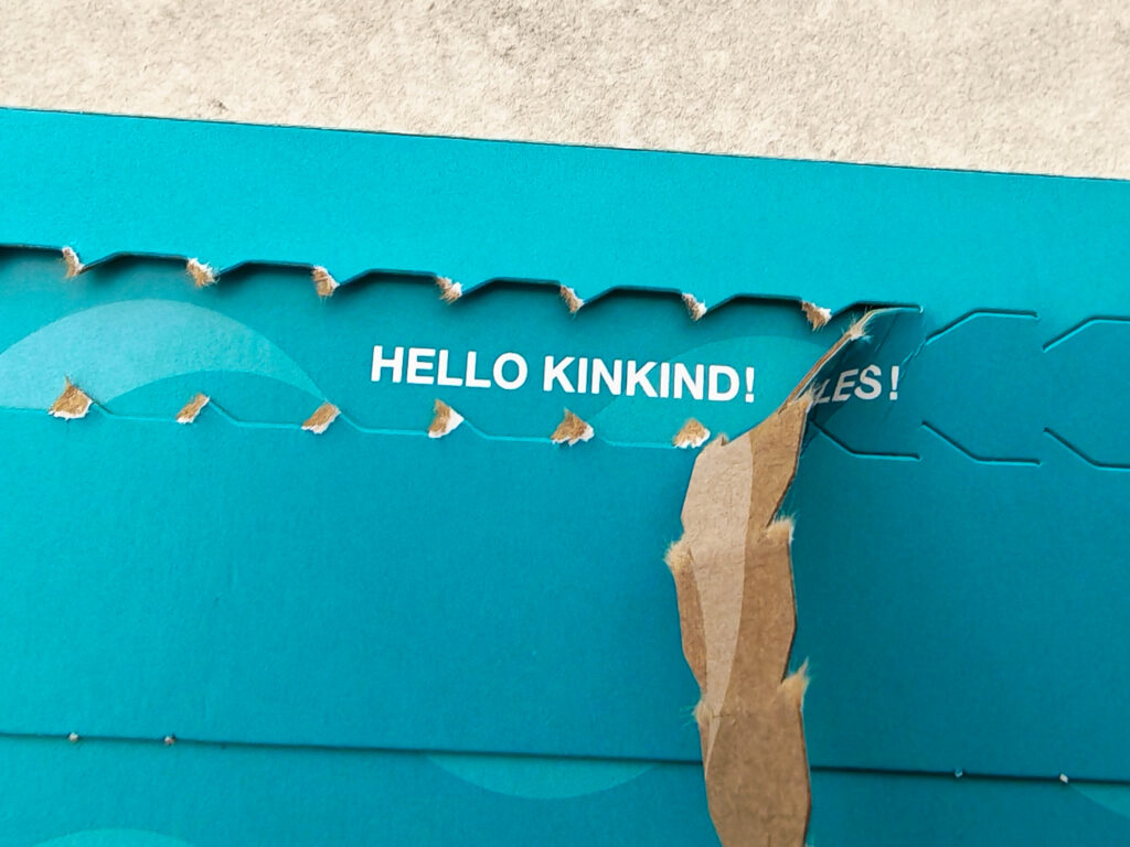 Torn cardboard envelope revealing HELLO KINKIND! text, highlighting blue packaging design with partial perforation.