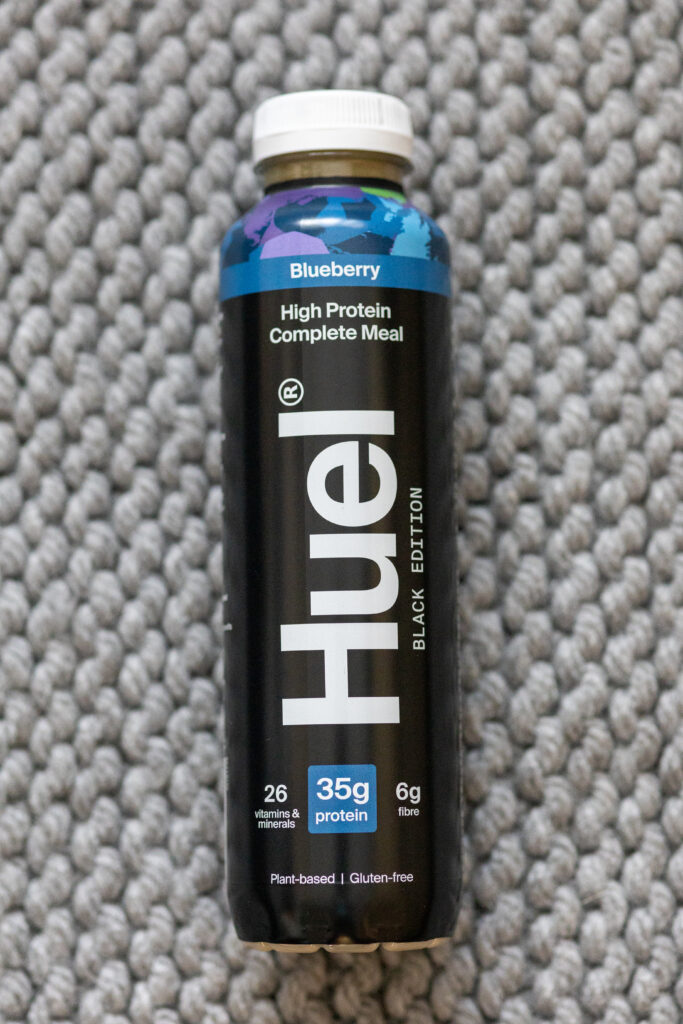 Huel Black Edition Blueberry bottle on textured grey surface, high protein, plant-based meal with 35g protein.