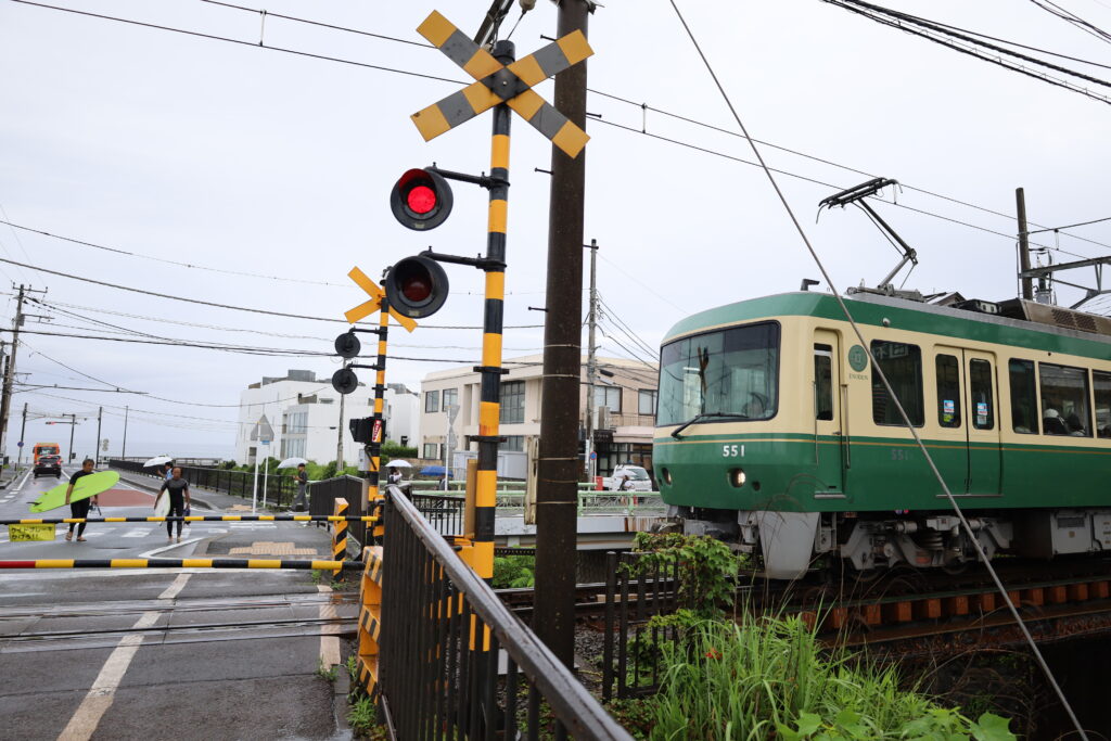 Green train at a railway crossing with surfers waiting, red signal shown, cloudy sky background.