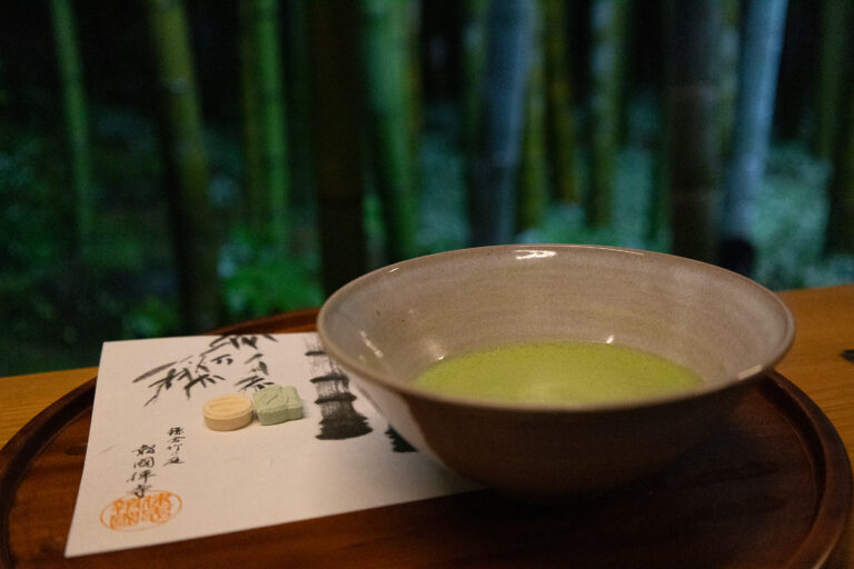 Ceramic bowl with green matcha tea on a tray, beside Japanese paper, set against a blurred bamboo background.