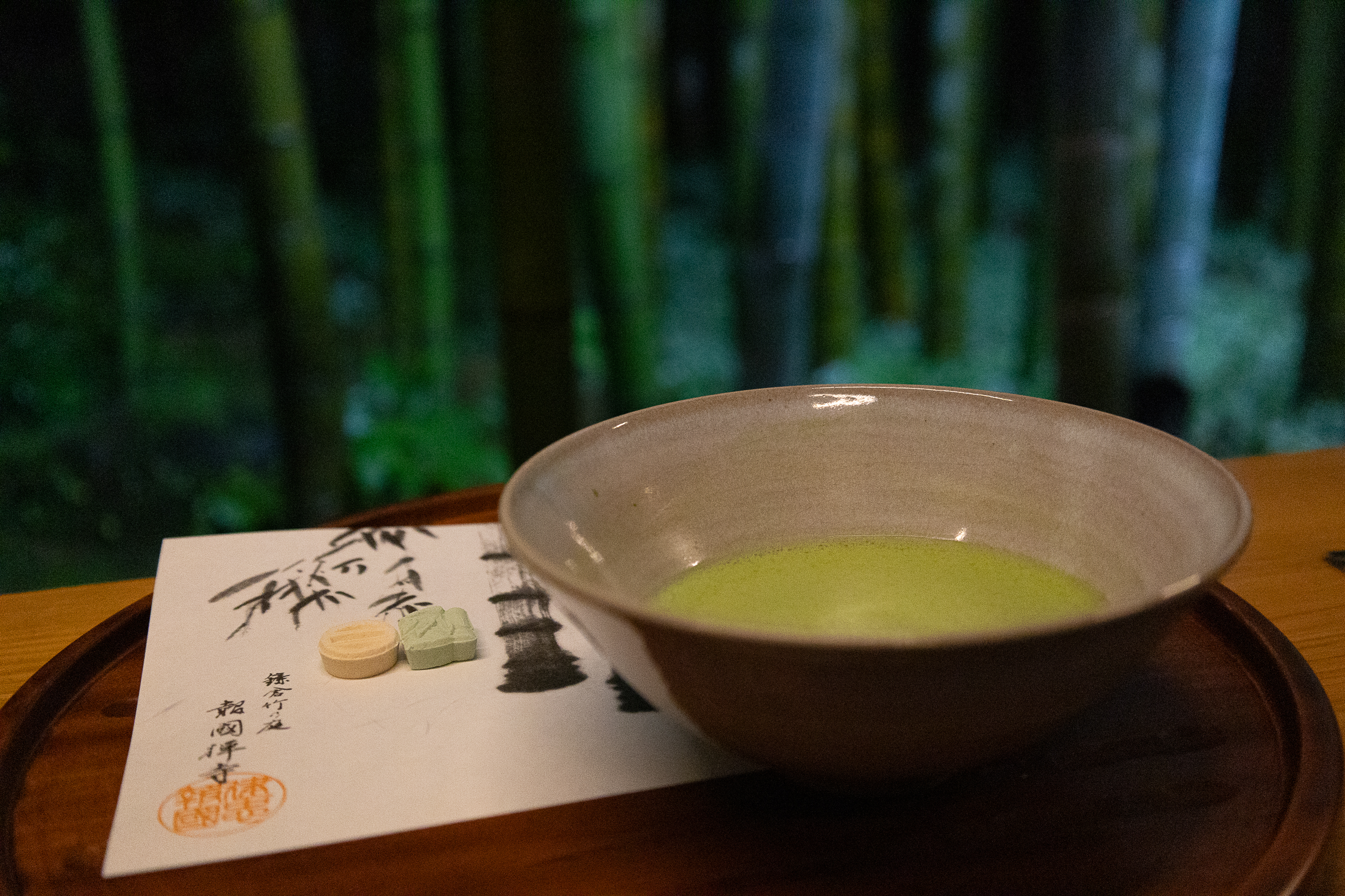 Ceramic bowl with green matcha tea on a tray, beside Japanese paper, set against a blurred bamboo background.