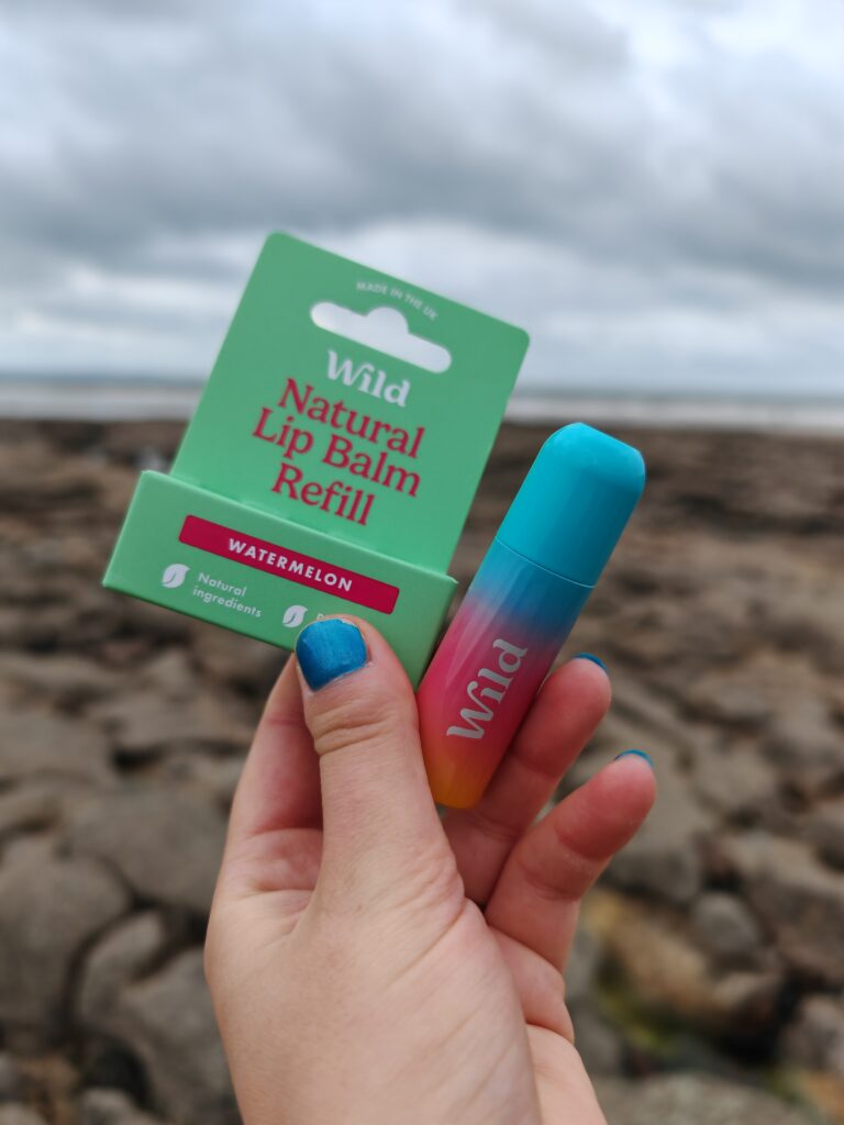Hand holding Wild natural lip balm refill, watermelon flavour, by the rocky seaside.