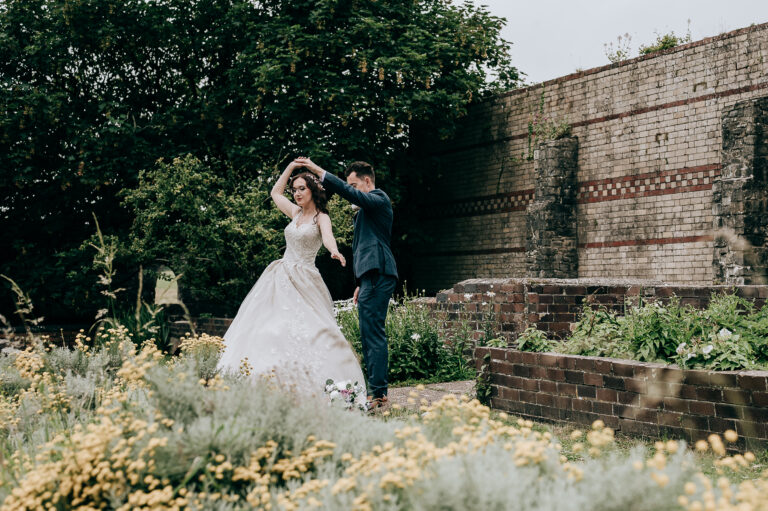 Bride and groom dancing joyfully in a garden setting with historic brick walls.