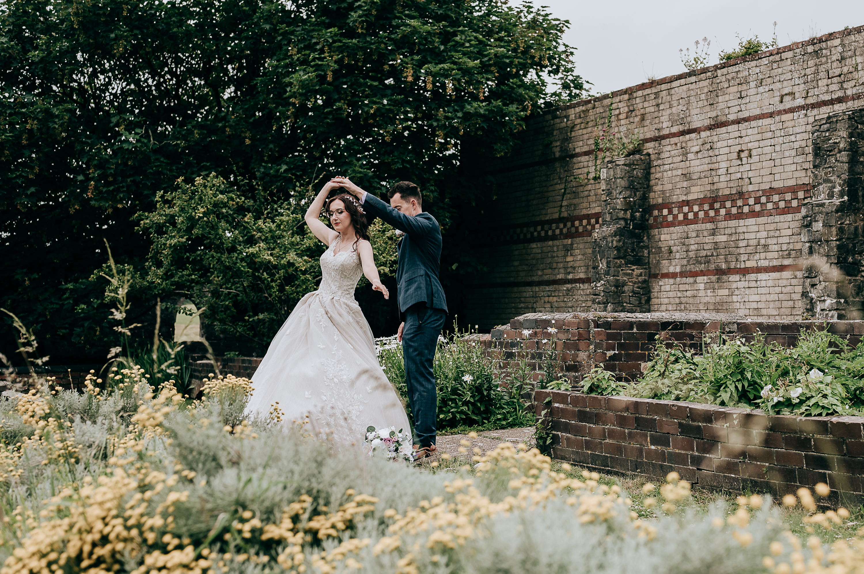 Bride and groom dancing joyfully in a garden setting with historic brick walls.