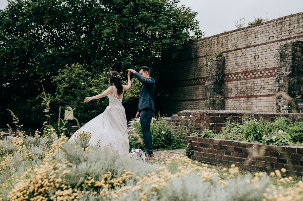 Bride and groom dancing in a garden with yellow flowers and an old brick wall background.