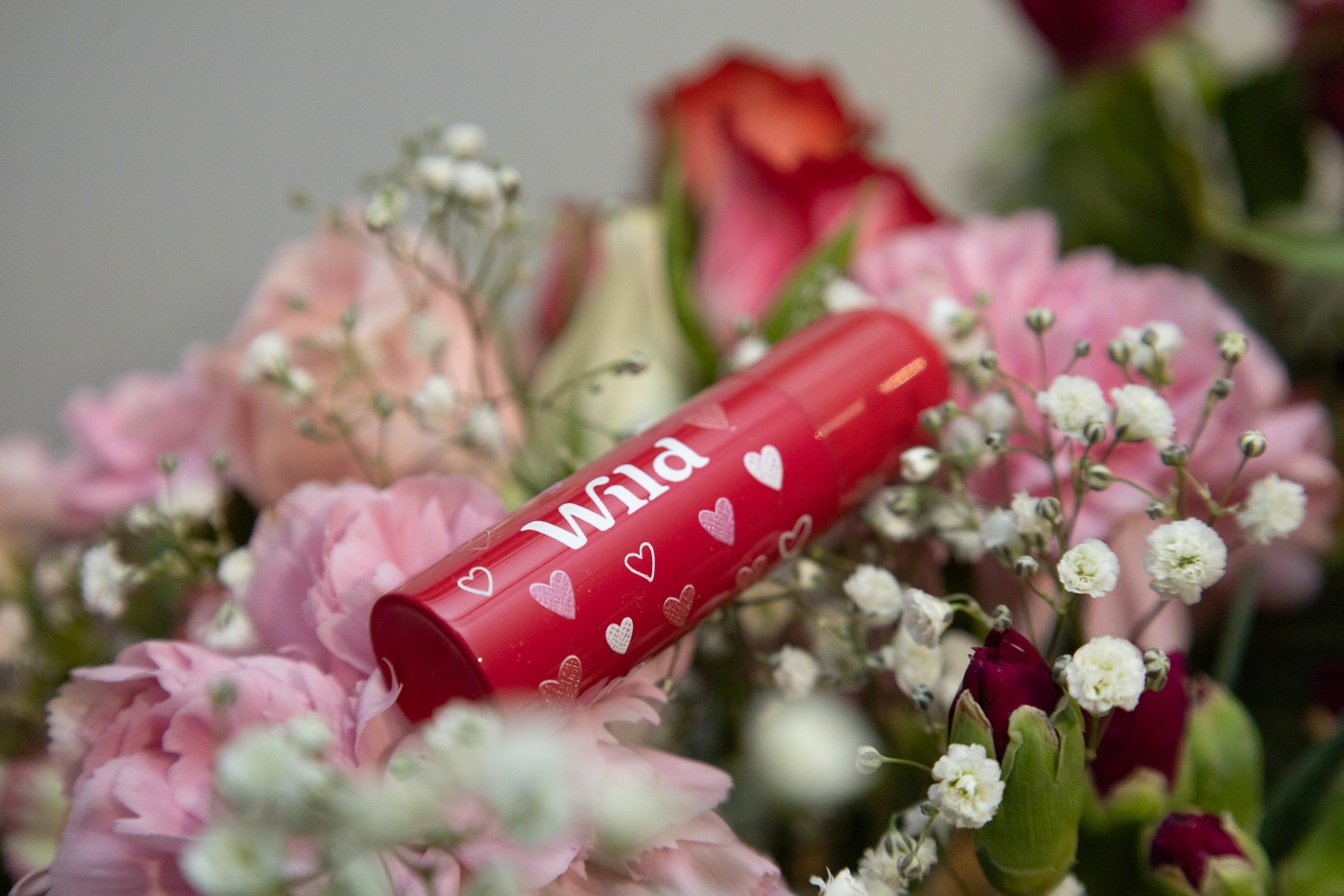 Red lipstick with heart pattern amidst pink and white flowers, promoting Wild beauty products in a floral setting.