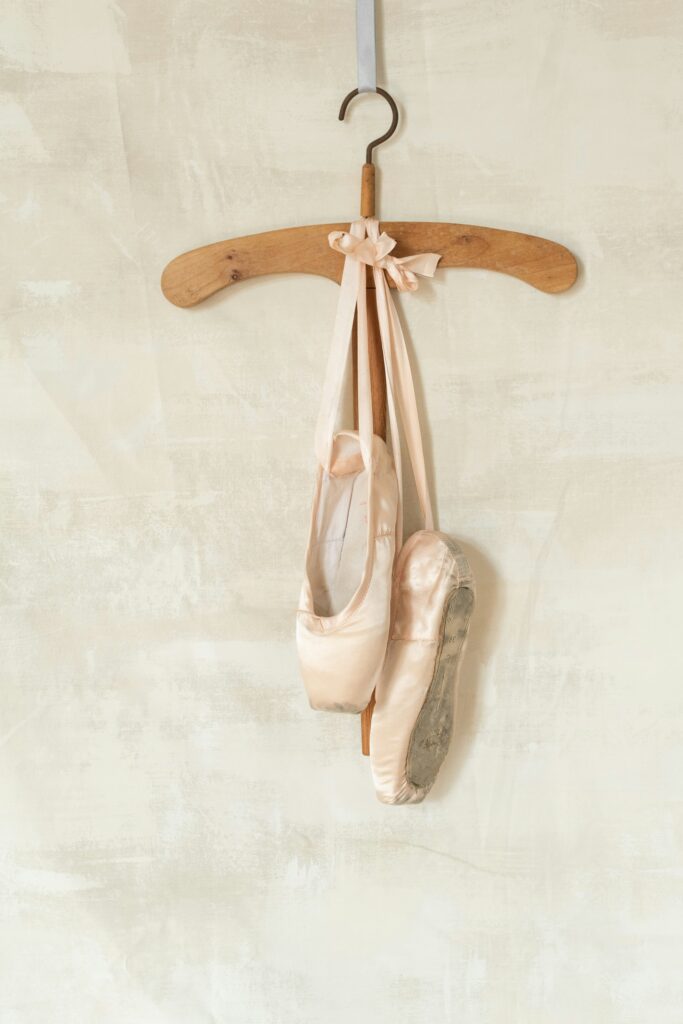 Pink ballet pointe shoes hanging on a wooden hanger against a beige textured wall, symbolising dance elegance.